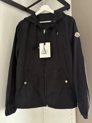 Svart Moncler vindjacka med huva - Svart vindjacka från Moncler med huva och dragkedja framtill. Jackan har två snedställda fickor med tryckknappar och Moncler-logga på vänster ärm. Materialet är lätt och vindtätt, perfekt för att hålla sig skyddad mot blåst. NFC, qr kod blipp allt funkar 