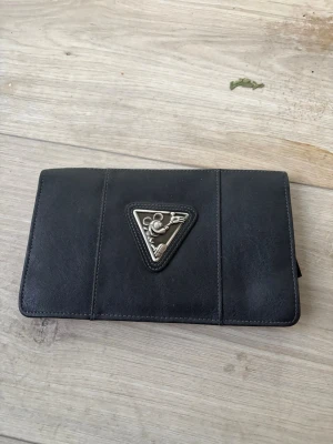 Svart plånbok med metalltriangel - Cool svart plånbok i läderimitation med ett metalltriangel-emblem med Musse Pigg på framsidan. Plånboken har en stilren och enkel design med diskreta sömmar och är perfekt för att hålla koll på kort och cash.