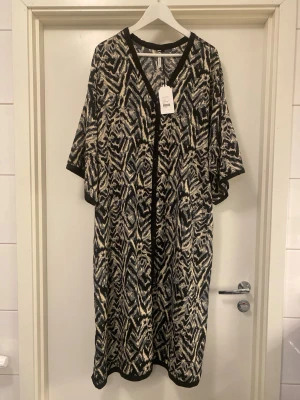 Långklänning med zebramönster - Snygg kaftan med svart, beige och vit abstrakt mönstring. Lång modell med vida ärmar och svarta kanter längs halsringning, ärmslut och slits. Luftigt och lätt material. Ny med prislapp!