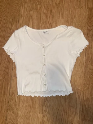 Vit ribbad topp med knappar Kappahl - Säljer en vit ribbad topp från Kappahl med korta ärmar och dekorativa knappar framtill. Toppen är croppad med volangkant vid ärmslut och nederkant.