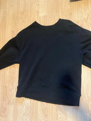 Svart sweatshirt utan huva - En enkel och stilren svart sweatshirt med rund hals och ribbade muddar. Tröjan har en klassisk passform och är gjord i mjukt material. Ingen huva och inget mönster.