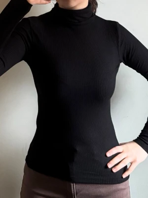 Svart ribbad polotopp - En svart, ribbad polotopp med långärmad design och tight passform. Perfekt för att skapa en clean och stilren look. Materialet är mjukt och stretchigt, vilket gör den bekväm att bära. Polokragen ger en klassisk touch och passar bra till olika outfits.