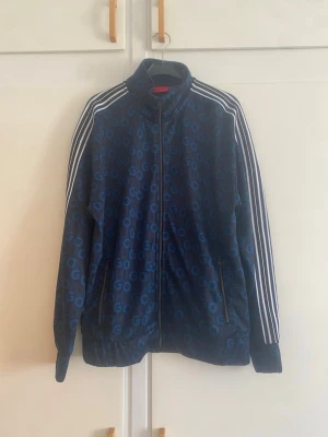 Mörkblå HUGO BOSS zip-tröja med mönster - Mörkblå zip-tröja från HUGO BOSS med ett coolt blått GO-mönster över hela plagget. Tröjan har klassiska vita ränder längs ärmarna, hög krage och två fickor med dragkedja. Perfekt för dig som gillar sportig och trendig stil.