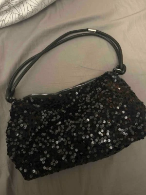 Svart axelväska med paljetter - Cool svart axelväska täckt med glittrande paljetter och ett stilrent handtag i svart material. Väskan har en rektangulär form och dragkedja upptill. Perfekt för dig som vill ha en trendig och unik accessoar som sticker ut.