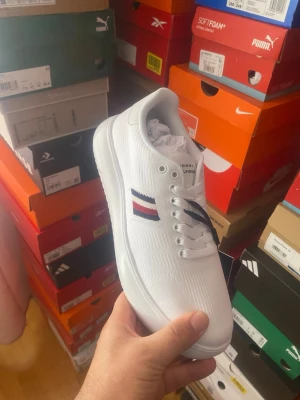 Vita sneakers från Tommy Hilfiger - Fräscha vita sneakers från Tommy Hilfiger med klassiska ränder i rött, vitt och blått på sidan. Lågt skaft, vita snören och diskret logga på plösen. Ovandelen är i textil och sulan är vit och stilren. Perfekta för en clean och modern look.