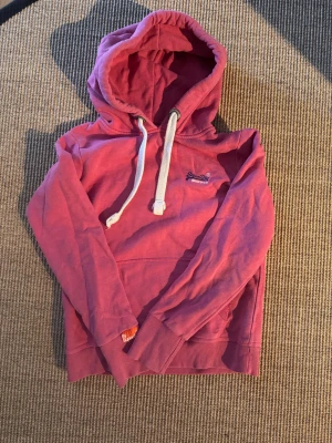 Ljus röd hoodie från Superdry - Säljer en snygg rosa hoodie från Superdry med vit dragsko i huvan och broderad logga på bröstet. Hoodien har en klassisk känguruficka framtill och ribbade muddar vid ärmar och nederkant. Perfekt för en avslappnad och trendig stil.