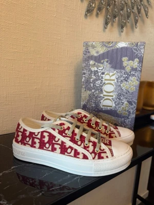 Dior sneakers Röd - Snygga låga sneakers från Dior med röd och vit monogramdesign. Materialet är canvas med gummisula och klassisk rund tå. Perfekt för dig som vill sticka ut med exklusiva sneakers.