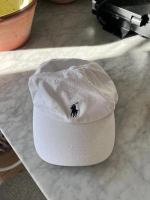 Vit keps - Säljer en vit keps med klassisk böjd skärm och broderad logga i svart framtill. Kepsen är tillverkad i bomull och har justerbar rem baktill för perfekt passform. Perfekt för en clean och sportig look.