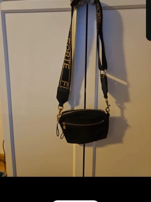 Svart axelväska  - Cool svart axelväska  med bred justerbar axelrem och guldfärgade detaljer. Väskan har en praktisk dragkedja framtill och är tillverkad i ett mjukt tygmaterial. Perfekt för att bära dina essentials och ge din outfit en trendig touch.