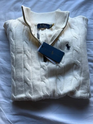 Ralph lauren stickad quarter zip  - Vit kabelstickad tröja från Polo Ralph Lauren med halv dragkedja och ribbad mönster. Den är helt oanvänd och skickad med taggar. Matrialet är perfekt och precis som den ska vara. Loggan är mer mörk bli men på bild ser den lite mer svart ut.