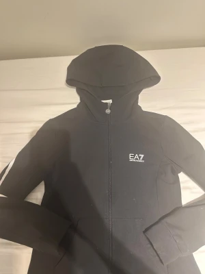 Svart hoodie från EA7 Emporio Armani - Snygg svart hoodie från EA7 Emporio Armani med dragkedja framtill och vit logga på bröstet. Klassisk huva och långa ärmar. Perfekt för en avslappnad och trendig stil. Tillverkad i mjukt material som känns skönt mot huden.