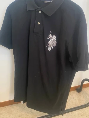 Svart Polo Assn pikétröja - Klassisk svart pikétröja från POLO Assn  med vit broderad polospelare på bröstet och siffran 3 på ärmen. Tröjan har korta ärmar, krage och knappar framtill. Perfekt för en stilren och sportig look.