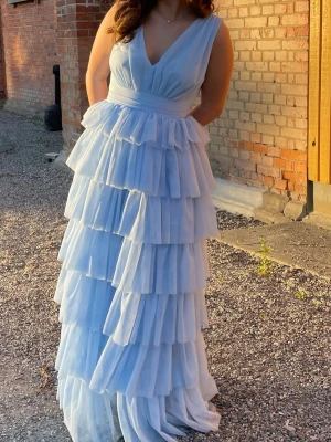 Ljusblå volangklänning med v-ringning - Säljer en lång ljusblå klänning med djup v-ringning och flera lager volanger i tyll. Klänningen har ärmlös design och markerad midja för en elegant siluett. Perfekt för bal eller festliga tillfällen där du vill sticka ut. Klänningen är använd en gång på bal, det finns lite fläckar från spraytan runt axlar, byst och rygg men ingen som syns när man har på sig den. Tror också det är utmärkt att försöka tvätta bort! 