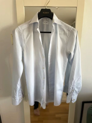 Ljusblå skjorta från Eton - Säljer en klassisk ljusblå skjorta från Eton med lång ärm och knappar framtill. Skjortan har en snygg krage och är tillverkad i bomull, perfekt för dig som gillar stilrena plagg. Passformen är normal och skjortan är enkel att matcha med olika outfits.