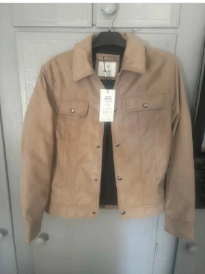 Beige mockajacka - Snygg beige mocka jacka med klassisk krage och raka linjer. Jackan har två bröstfickor med lock och silverfärgade knappar framtill. Perfekt för dig som vill ha en stilren och enkel look. Passar till det mesta och är lätt att matcha. Köpte svart o beige så väljer att sälja denna helt nya som är super till vår och sommar. Passar även S. Använd köp nu