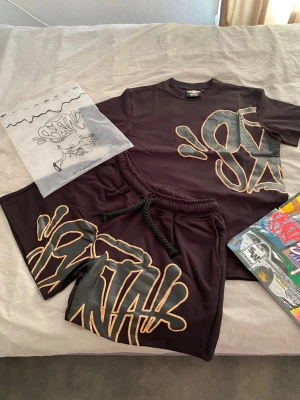 Svart set från Syna world  - Säljer ett svart set från Syna world med matchande t-shirt och shorts. Båda plaggen har stort graffiti-inspirerat tryck i grått och beige på framsidan. T-shirten har rund hals och korta ärmar, shortsen har resårmidja med snörning. Perfekt för en avslappnad streetwear-look.