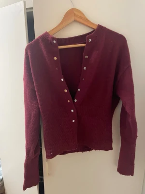 Vinröd kofta med vita knappar - Mysig vinröd kofta med ribbstickad nederkant och vita knappar framtill. Koftan har lång ärm och är tillverkad i ett mjukt stickat material som känns skönt mot huden. Perfekt att slänga på sig när det är lite kyligt.