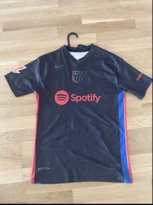 FC Barcelona matchtröja svart Nike - Snygg svart FC Barcelona matchtröja från Nike med röda och blå detaljer på sidorna. Tryck med Spotify-logga på bröstet och Lamine Yamal 19 på ryggen. Tillverkad i lätt och ventilerande material, perfekt för fotboll eller träning. Storleken är M men skulle kunna funka som S.