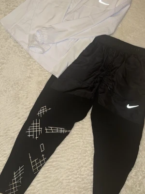 Nike Tracksuit  - Svarta löparbyxor från Nike med vita grafiska mönster på benen och reflekterande logga. Byxorna har elastisk midja och är tillverkade i ett lätt och stretchigt material som passar för löpning och träning. Medföljer med en vit Nike vindjacka i nytt skick inget fel elle annat på Tracksuiten storlek M 