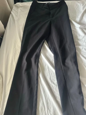 Svarta kostymbyxor med pressveck - Snygga svarta kostymbyxor med raka ben och pressveck. Byxorna har hällor för skärp och stängs med dragkedja och knapp. Perfekta för en clean och stilren look.