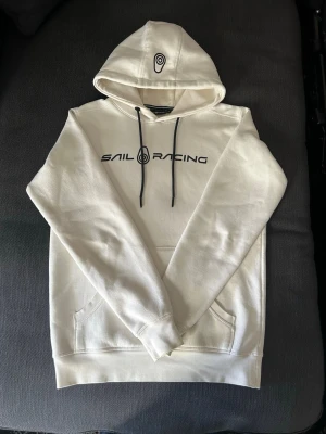 Vit hoodie  Sail Racing - Vit hoodie från Sail Racing med svart logga och text på bröstet. Hoodien har svarta dragsnören, känguruficka och en broderad logga på huvan. Tillverkad i mjukt bomullsmaterial och har ribbade muddar vid ärmslut och nederkant.