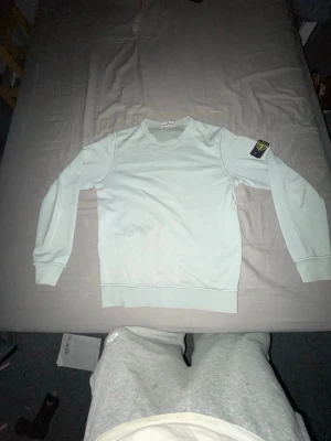 Ljusblå sweatshirt från Stone Island - Ljusblå sweatshirt från Stone Island med klassisk rund hals och ribbade muddar. Ikonisk logopatch på vänster ärm. Perfekt för dig som gillar streetwear och vill ha en clean look. Materialet är mjukt och bekvämt, passar till jeans eller joggers.
