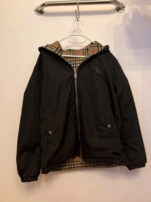 Svart vindjacka med rutigt foder - Svart vindjacka med dragkedja och huva. Insidan har klassiskt beige-rutigt foder. Jackan har två fickor framtill med tryckknappar och elastiska muddar vid ärmslut. Perfekt för blåsiga dagar och enkel att matcha med olika outfits.