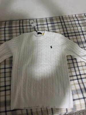 Vit kabelstickad tröja Polo Ralph Lauren - Kabelstickad vit tröja från Polo Ralph Lauren med rund halsringning och den klassiska broderade loggan på bröstet. Tröjan har långa ärmar och ribbade muddar vid ärmslut och nederkant. Perfekt för en stilren och tidlös look.