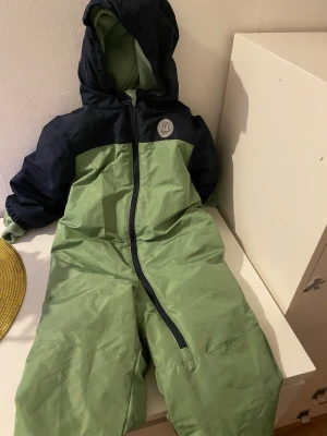 Grön täckbyxa med huva från M - Säljer ett par gröna täckbyxor med mörkblå detaljer och huva från märket M. Dragkedja framtill och reflexdetalj på bröstet. Perfekt för kalla dagar och utomhusaktiviteter. Materialet är slitstarkt och vindtätt.