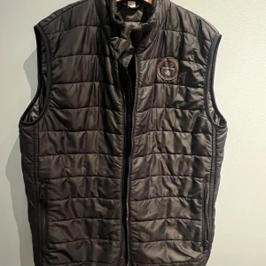 Napapijri vadderad väst – svart | Strl 152 (12 år) - Snygg och praktisk vadderad väst från Napapijri i svart. Perfekt som lager-på-lager under vår och höst eller över hoodie/sweater. Klassisk quiltad design med dragkedja framtill och fickor med dragkedja. Ikonisk Napapijri-logga på bröstet. Märke: Napapijri Storlek: 152 / 12 år Färg: Svart Modell: Vadderad väst Skick: Bra använt skick, inga större anmärkningar Tillverkad i Marocko En tidlös och användbar väst som passar till det mesta