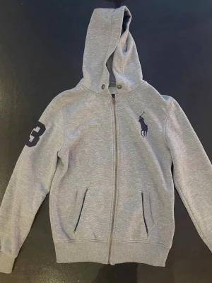 Grå hoodie från Polo Ralph Lauren - Snygg grå hoodie med dragkedja från Polo Ralph Lauren. Stor logga i mörkblått på bröstet och en stor siffra 3 på ärmen. Två fickor fram och ribbade muddar. Tillverkad i mjuk bomullsblandning och har en klassisk huva.
