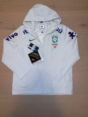 Vit/beige Nike Windbreaker Brazil Gore-Tex - Snygg vit/beige windbreaker från Nike med Brazil-logga och flera sponsortryck på ärmar och bröst. Jackan har huva, hel dragkedja och är tillverkad i GORE-TEX-material som skyddar mot vind och regn. Perfekt för dig som gillar sportig stil och vill sticka ut.