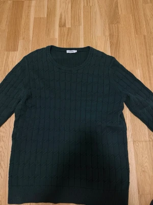 Mörkgrön stickad tröja J.lindberg - Mörkgrön stickad tröja från J.Lindberg med rund halsringning och ett diskret kabelstickat mönster över hela tröjan. Långa ärmar och ribbade muddar vid ärmslut och nederkant. Perfekt för kyliga dagar och enkel att matcha.
