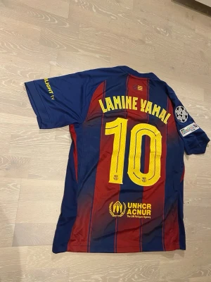 Barcelona matchtröja Lamine Yamal #10 - Säljer en officiell FC Barcelona matchtröja med Lamine Yamal och nummer 10 på ryggen. Tröjan har klassiska blå och röda vertikala ränder, gula tryck och Champions League-märke på ärmen. Tillverkad i lätt och ventilerande material, perfekt för fotboll.