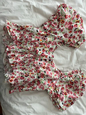 Blommig croppad blus med puffärm - Supersöt blommig croppad blus med puffärmar och smockad midja. Toppen har volangkant nedtill och ett färgglatt mönster i rosa, rött, orange och grönt på vit botten.