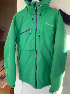 Grön skidjacka från Peak Performance - Snygg grön skidjacka från Peak Performance med huva, flera dragkedjeförsedda fickor och justerbara detaljer. Jackan har blå dragkedjor och logga på bröstet. Gore tex