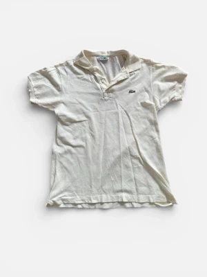 Gul piké från Lacoste  - Klassisk Gul pikétröja från Lacoste med korta ärmar och krage. Tröjan har två knappar vid halsen och den ikoniska gröna krokodilloggan broderad på bröstet. Den har sytts under vänstra armen men det syns inte när man har på sig pikén.Tillverkad i mjuk bomull som känns skön mot huden. Perfekt för en clean och sportig look.