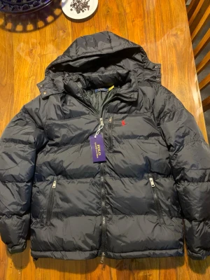  Ralph Lauren jacka  - Svart dunjacka från Polo Ralph Lauren med huva och två dragkedjeförsedda fickor. Jackan har det klassiska röda Polo-logot broderat på bröstet och en hög krage. Materialet är glansigt och jackan har en puffig, quiltad design. Perfekt för kalla vinterdagar.