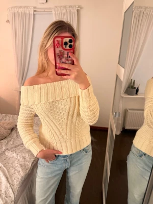 Off shoulder stickad tröja i creme - Trendig off shoulder stickad tröja i cremefärg med bred ribbad kant vid axlarna och snyggt kabelstickat mönster. Tröjan har lång ärm och är gjord i ett mjukt material som känns skönt mot huden. Perfekt för dig som vill ha en stilren men ändå cozy look.