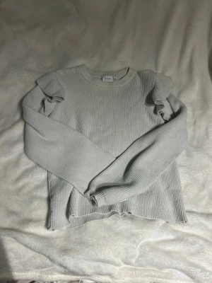 Vit tröja med volang - Supersnygg vit stickad tröja från Vila med ribbad struktur och volangdetaljer på axlarna. Tröjan har rund halsringning och långa ärmar. Perfekt för dig som gillar en enkel men trendig look.