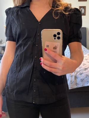 Svart kortärmad skjorta med puffärm - Stilren svart skjorta med korta puffärmar och v-ringning. Skjortan har knappar framtill och en lätt figurnära passform. Perfekt för dig som gillar en enkel men trendig look. Saknar ett knäppe till knappen längst ner men inget som syns.