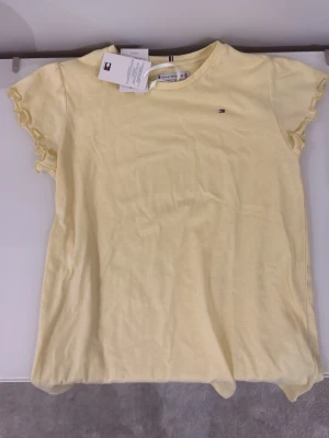 Gul t-shirt från Tommy Hilfiger - Snygg gul t-shirt från Tommy Hilfiger med liten logga på bröstet. T-shirten har korta ärmar med volangkant och rund halsringning. Tillverkad i mjuk bomull, perfekt för en avslappnad stil.