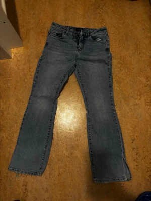 Blå bootcut jeans med slits - Säljer ett par blå bootcut jeans i klassisk denim med slits nedtill på benen. Jeansen har fem fickor och rå kant längst ner för en trendig look. Perfekta för dig som gillar en avslappnad men ändå stilsäker stil. Står ingen storlek men skulle säga s/36