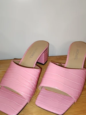 Rosa sandaler med blockklack från Monki - Snygga rosa sandaler från Monki med breda band och croco-mönstrad yta. De har fyrkantig tå och stabil blockklack, vilket ger en trendig look. Materialet är konstläder och sulan är svart. Perfekta för dig som vill sticka ut lite extra.