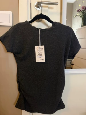 Mörkgrå ribbad topp med snörning - Mörkgrå topp i mjukt material. Kortärmad modell med rund halsringning och snygg snörningsdetalj i sidan som ger en cool twist. Perfekt till jeans eller kjol för en avslappnad look.