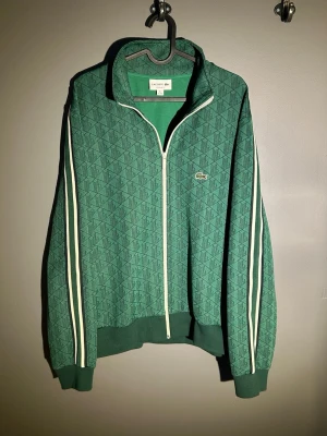 Grön zip hoodie från Lacoste - Grön hoodie från Lacoste med hel dragkedja och diskret mönster i tyget. Klassiska vita ränder längs ärmarna och broderad Lacoste-logga på bröstet. Ribbstickade muddar och krage. Tillverkad i mjukt material som känns skönt mot huden. Storlek XXL men passar bäst L/XL. Kvitto finns