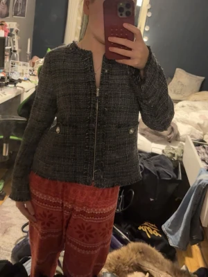 Svart tweedkavaj med dragkedja - Stilren svart tweedkavaj med dragkedja framtill och dekorativa fickor med knappar. Kavajen har en rak passform och råa kanter vid halsringning och ärmslut. Perfekt för en trendig och klassisk look.