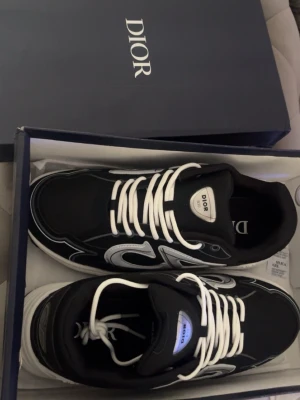 Dior b30  - Säljer ett par svarta Dior sneakers med vita och silverfärgade detaljer samt vita skosnören. Skorna har en chunky sula och tydlig Dior-logga på plösen. Materialet är en mix av mesh och läder för en modern och sportig look. Kommer i originalkartong.