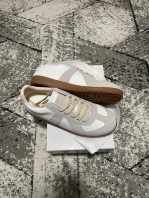 Vita skor med bruna sulor från maison margiela  -  Margiela Replica sneakers i vitt och beige med ovandel i läder och mocka, samt klassisk gummisula i brunt. Snygg retrostil med låga snitt och neutrala färger som passar till många outfits. Ikonisk design med diskreta detaljer och bekväm passform. Priset kan alltid diskuteras, jag fick den som present av en kusin och den va för liten så därför jag säljer den 