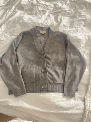 Grå kofta med stora knappar - Mysig grå kofta med v-ringning och tre stora knappar framtill. Två praktiska fickor på framsidan och ribbade muddar vid ärmslut och nederkant. Perfekt för lager på lager och enkel att matcha med olika outfits.